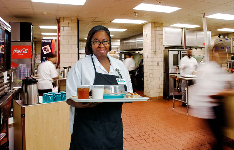 A smiling food service employee in an apron and hair net, holding a tray of food in the middle of the large hospital cafeteria kitchen.