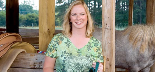 Lexington Medical patient Kimberly Williamson wears a green floral shirt and stands smiling in a stable holding her horse's reins.