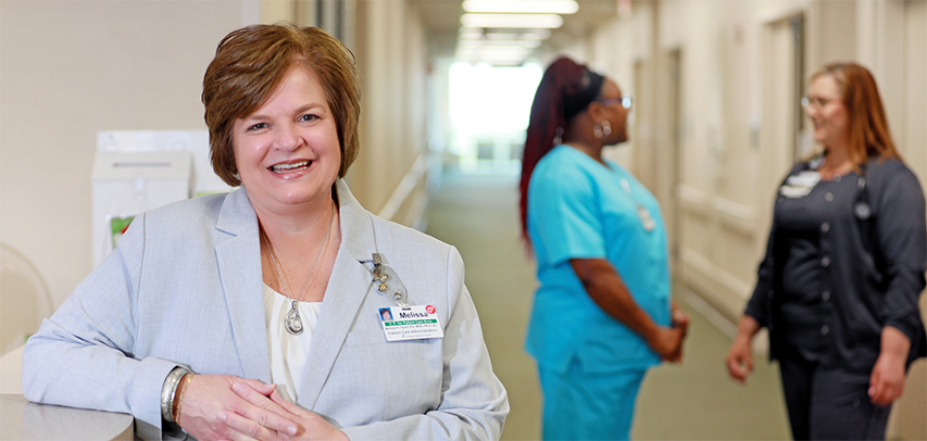 Chief Nursing Officer Melissa Taylor leaning on the reception counter, smiling and wearing a blazer, with two nurses in blue scrubs chatting behind her.