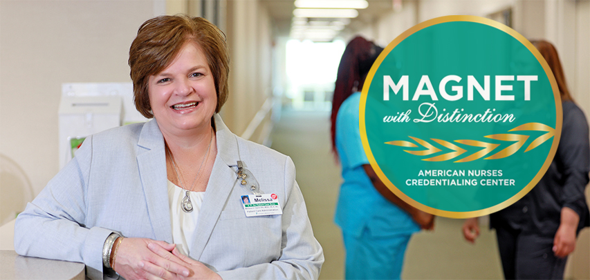 Chief Nursing Officer Melissa Taylor leaning on the reception counter, smiling and wearing a blazer, with two nurses in blue scrubs chatting behind her.