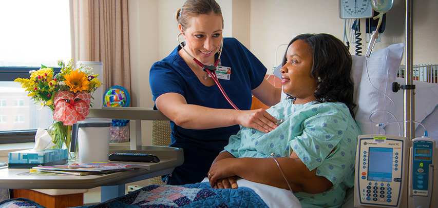 A nurse checking a patient's heartbeat with a stethoscope, both smiling, with flowers and balloons on the tray table next to them.