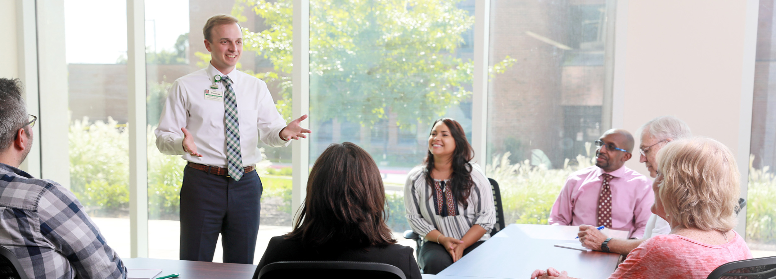 Community manager Thomas Tafel holds a roundtable discussion in a sunny room with large windows showing the tree-lined LMC campus.