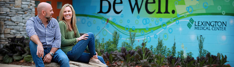 A smiling man and woman sitting in front of a wall with Lexington Medical Center’s logo and their slogan, 'Be Well.'