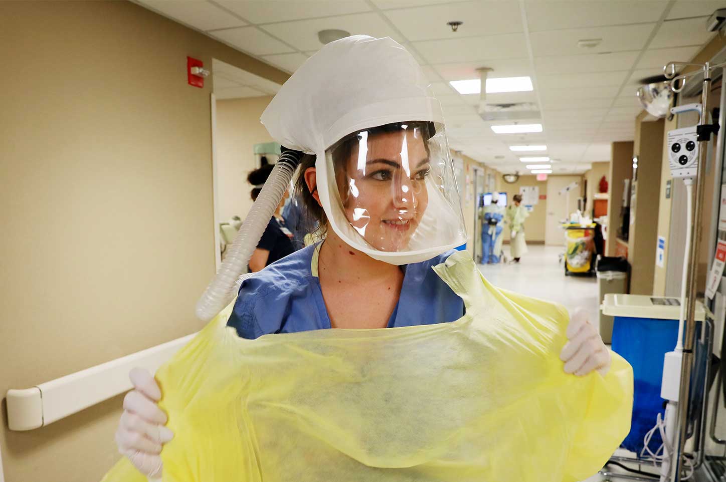 A frontline worker wearing full scrubs and face shield takes off a disposable hospital gown in a busy hallway.