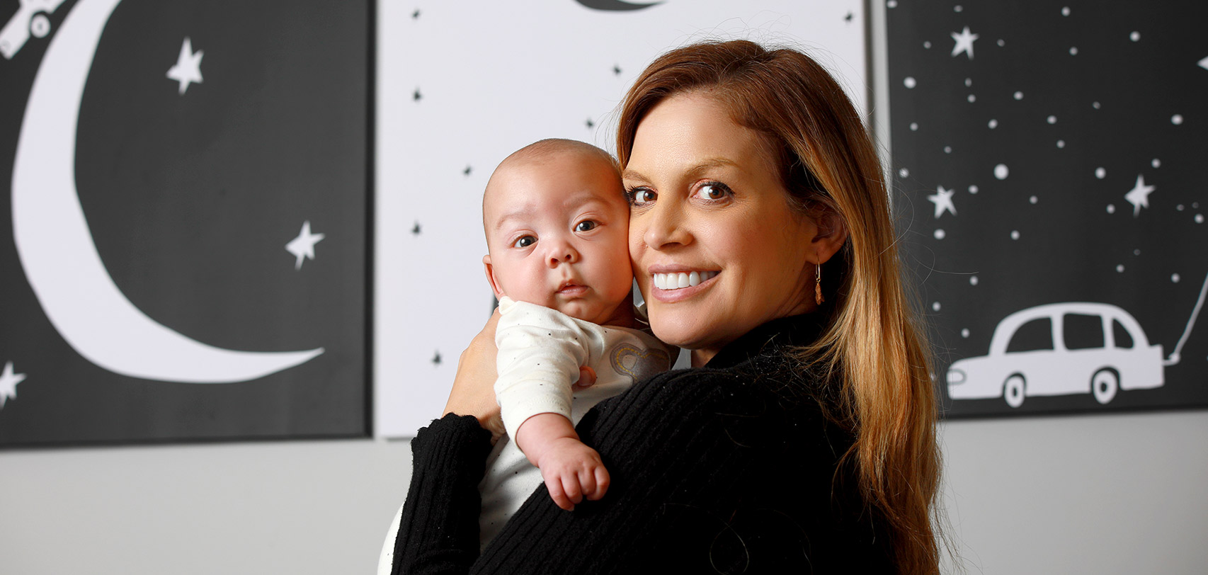 A mother holding her child in front of a series of black & white moon and star-themed paintings.