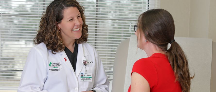 A female doctor in a white lab coat smiles warmly at a young patient in an exam room.