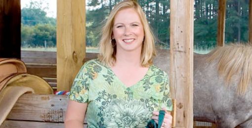 Lexington Medical patient Kimberly Williamson wears a green floral shirt and stands smiling in a stable holding her horse's reins.