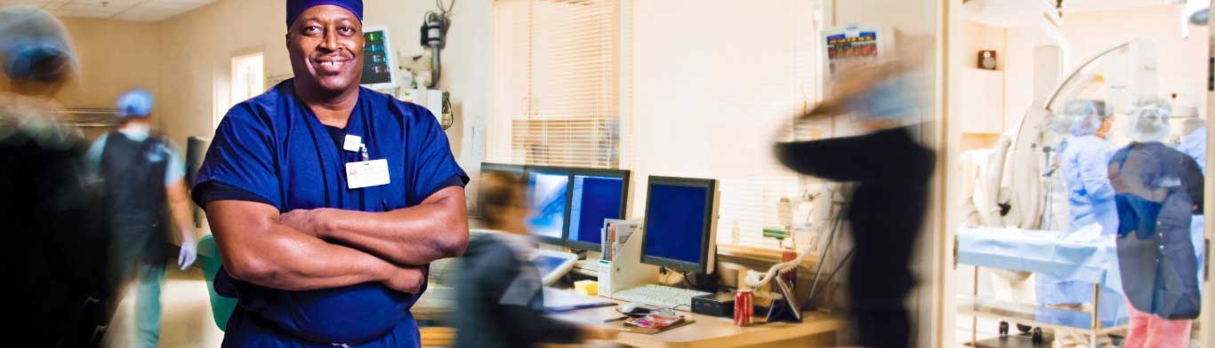 On a busy hospital floor, a care provider stands smiling with crossed arms in dark blue scrubs as nurses move around him in a blur of motion.