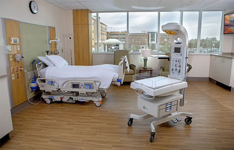 The inside of a spacious delivery suite with wood floors, large windows letting in lots of light, a hospital bed, a newborn crib, two arm chairs, and a large TV.