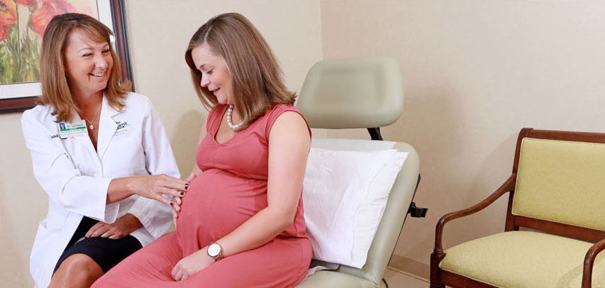 A female LMC doctor and pregnant patient smiling in a warm examination room. The patient has her hand on her baby bump while. The doctor has fingertips on the patient’s stomach, as if explaining something about the baby. 