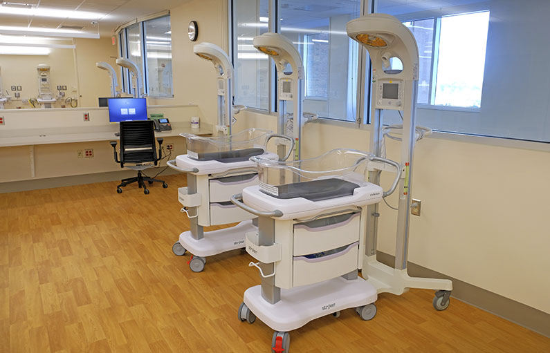 Newborn cribs in front of several windows in a spacious yet enclosed room with wood floors.