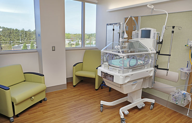 A special care newborn crib in a private special care nursery room with two green armchairs under large windows letting in lots of light and a view.
