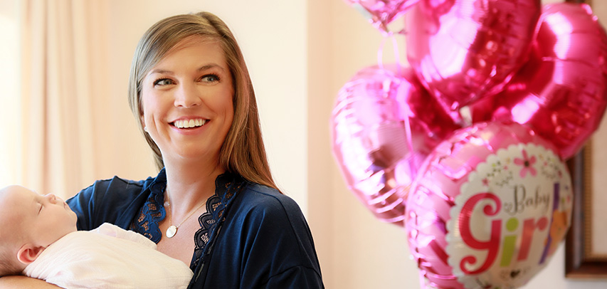 A patient smiling and holding her newborn baby swaddled in a blanket in her arms with a cluster of pink and white mylar “Baby Girl” balloons in the background.