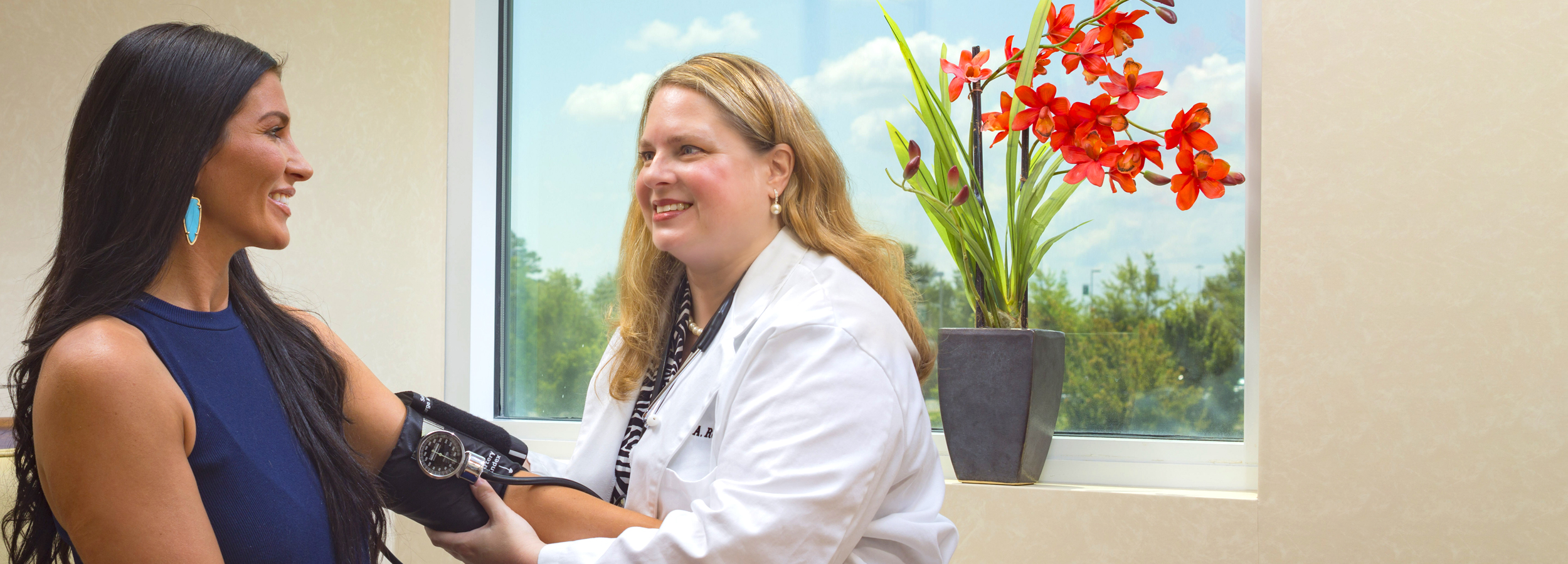 A female LMC doctor wearing a lab coat over a cheerful floral shirt holding paperwork and talking with a female patient in a warm siting room with wood, art and cushioned chairs.