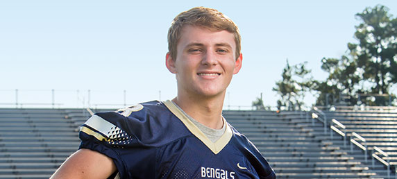 Sports injury patient Eli Ailshie in a football uniform holding his helmet under his arm on a football field.