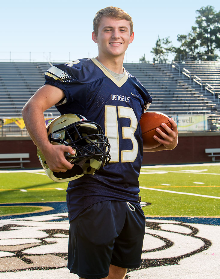Sports injury patient Eli Ailshie in a football uniform holding his helmet under his arm on a football field.