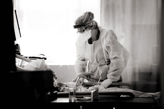 A care provider in full PPE and face shield standing beside an elderly patient’s bed, leaning in to listen, with medical equipment silhouetted in the foreground.